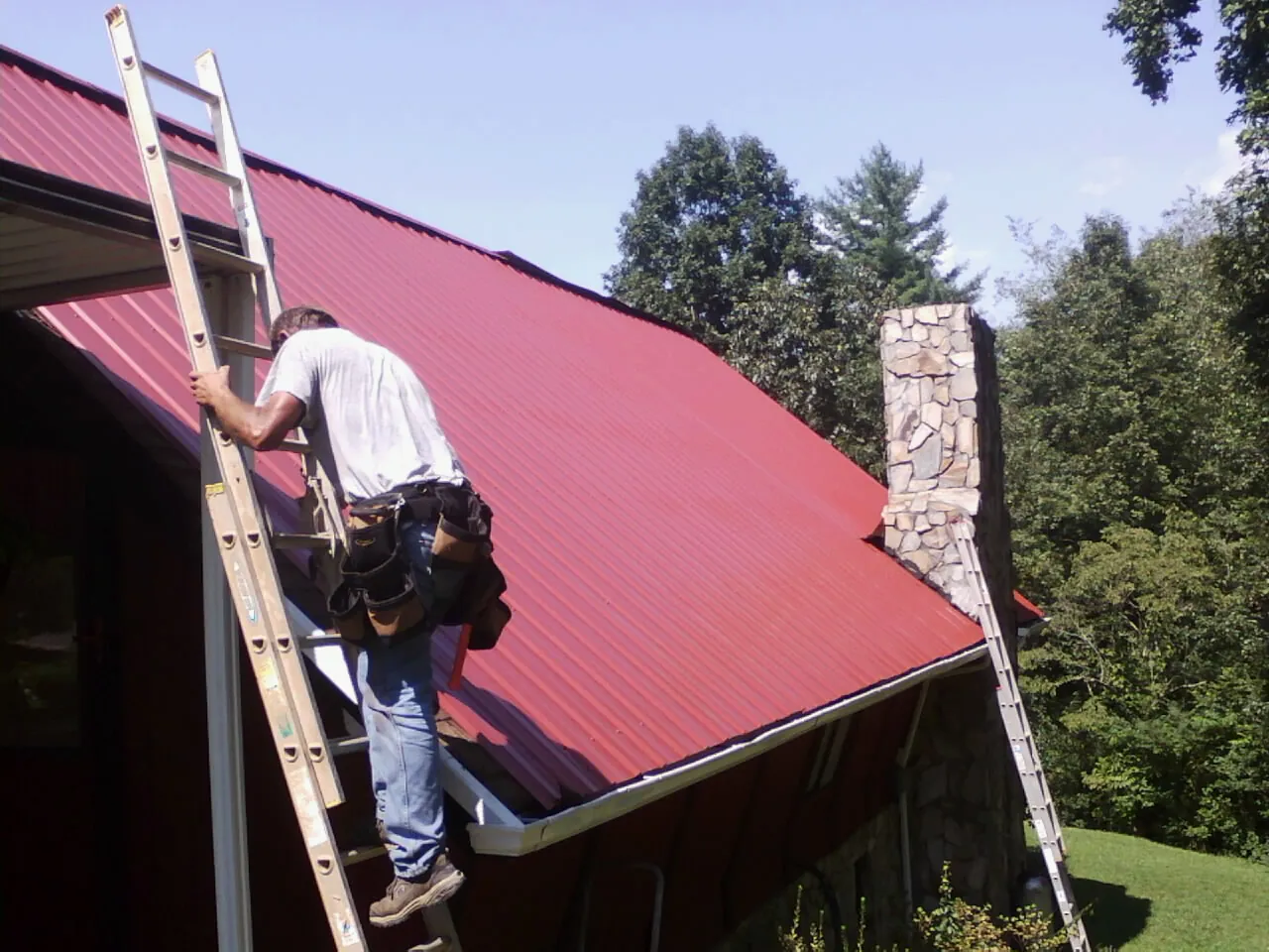 Roofer on a ladder beside a red metal roof and stone chimney — Soffit & Fascia Repair by Roofing Brothers in Corbett